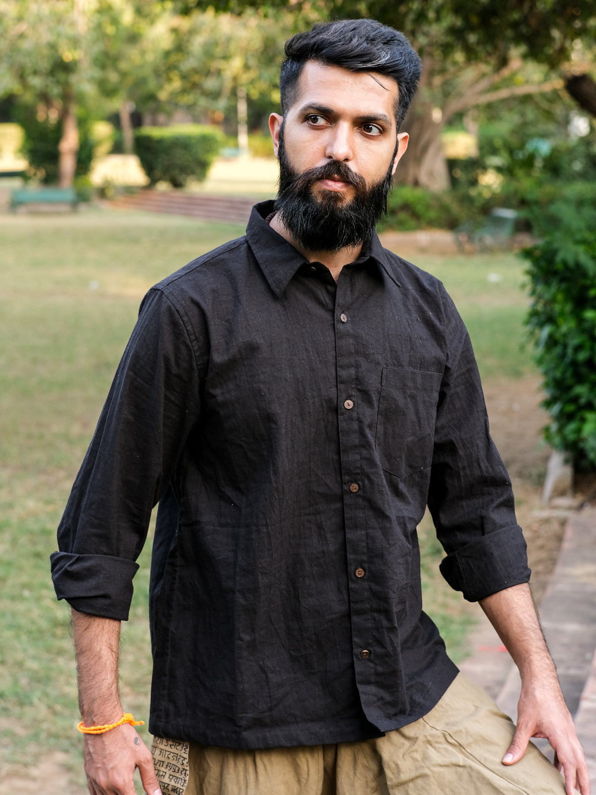 A bearded man with short dark hair stands outdoors in a park with greenery in the background, wearing a Bhadra Shirt made from premium fabric.