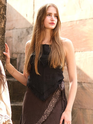 A woman with long, light brown hair stands outdoors by a tree, wearing the Chiara Crop Top—a black sleeveless boho style—paired with a brown skirt with a patterned sash, looking off into the distance.