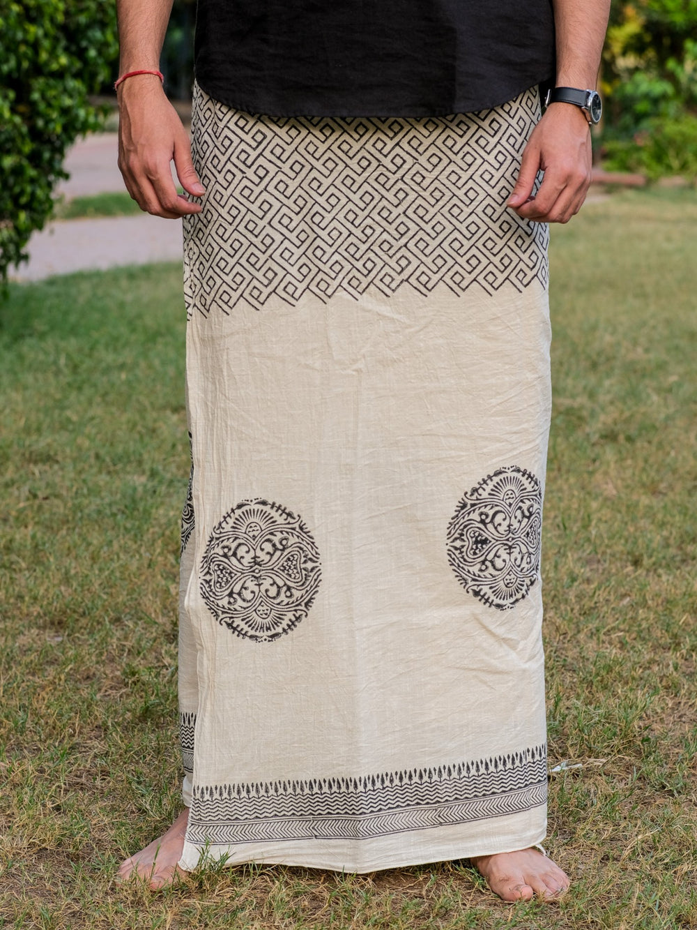 A person stands barefoot on grass, wearing an Aastha Dhoti in cream cotton with black geometric and circular patterns, their hands relaxed at their sides.