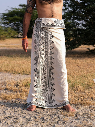 A person stands outdoors on dry grass and sand, wearing a Vyom Dhoti with black and white geometric block print patterns, set against a backdrop of trees and a clear sky.