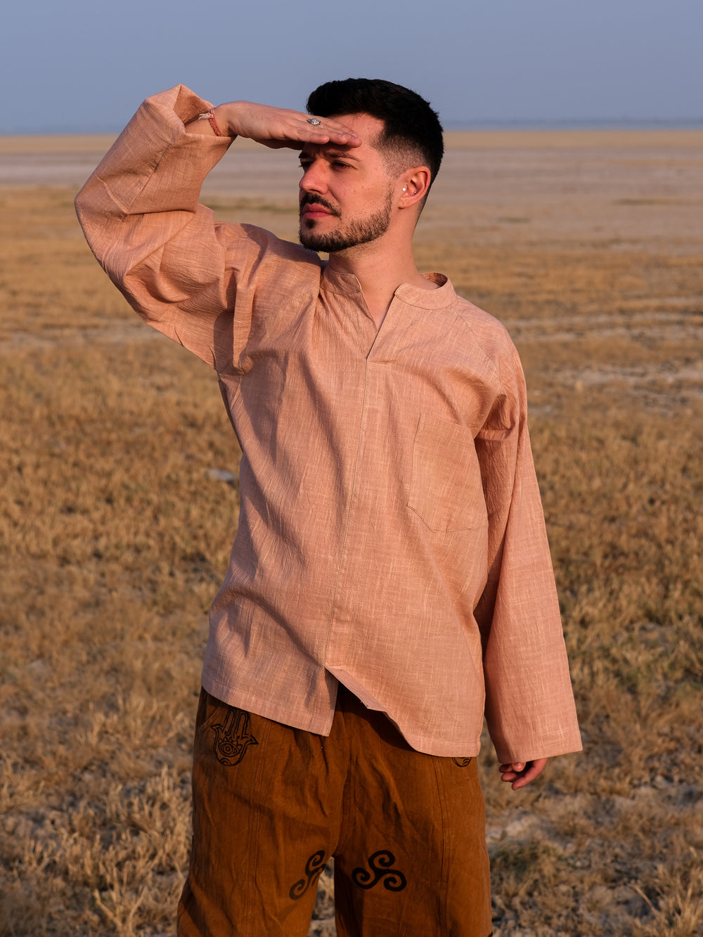 Wearing the Shlok Kurta in beige, a man stands in a dry, grassy field, shielding his eyes as he gazes into the distance on a sunny day. The loose fit and light hue of his outfit enhance his effortless boho style as he takes in the sunlit landscape.