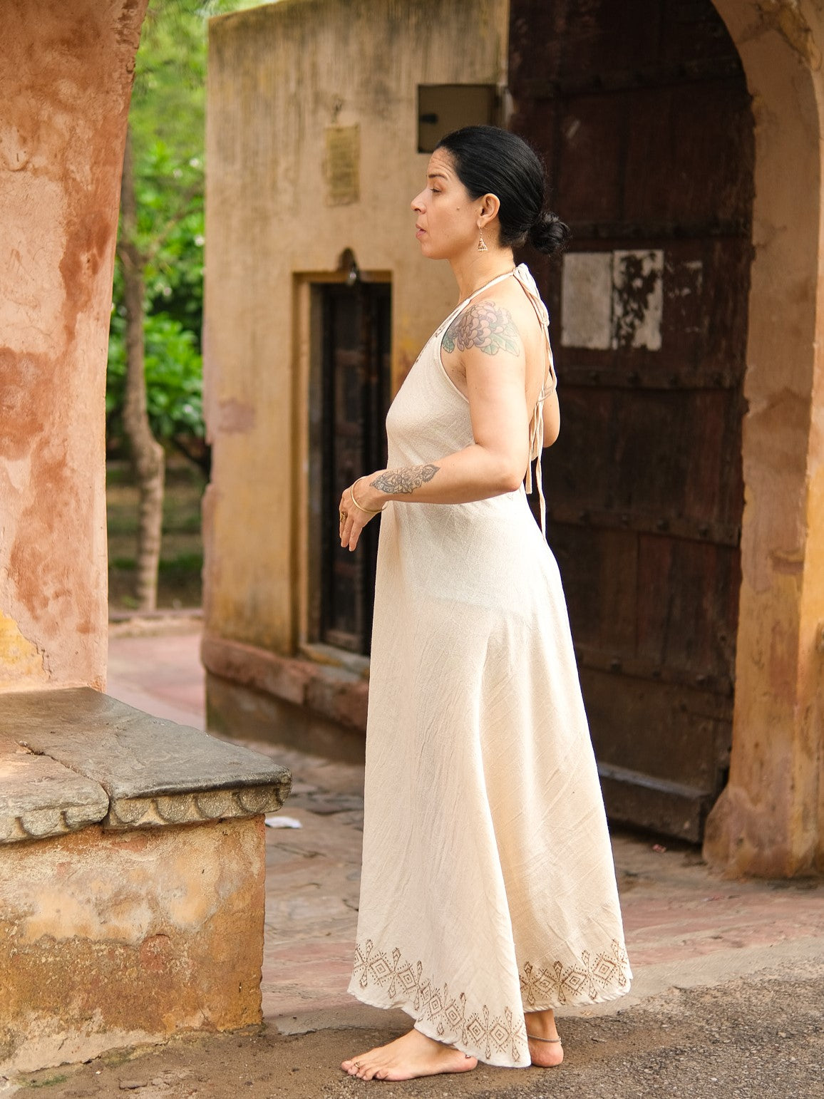 A woman with tattoos stands barefoot near an old rustic building with arched doorways and weathered walls, looking thoughtful. She wears a sleeveless cream Hanvi maxi dress made from sustainable cotton. Greenery can be seen in the background.