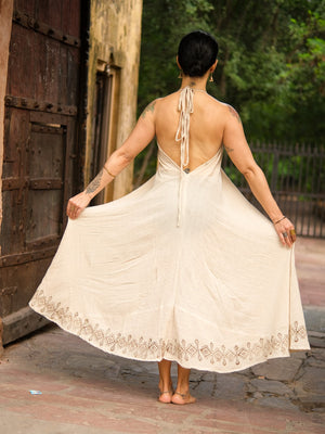 A barefoot person stands outdoors with their back to the camera, holding out the sides of the Hanvi maxi dress, crafted in cream sustainable cotton with decorative trim and subtle block print details, revealing their tattoos and an old building in the background.