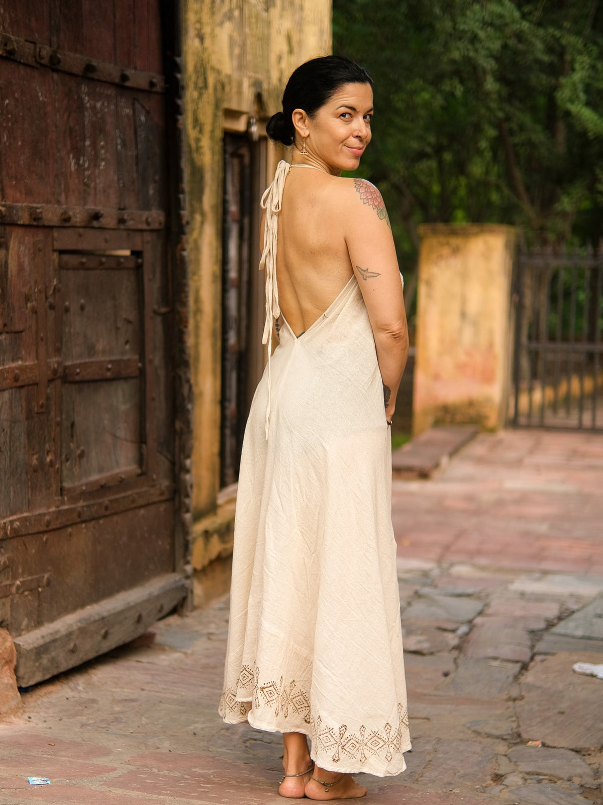 A woman, barefoot on a stone path and smiling over her shoulder, wears the Hanvi maxi dress—a cream, backless design crafted from sustainable cotton with delicate block print details. The dress accentuates her tattoos and dark hair tied back, while a rustic wooden door and lush greenery provide an inviting backdrop.