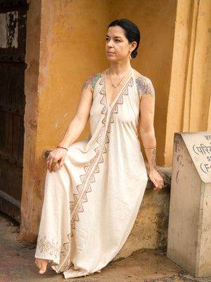 A woman with tattoos sits on a ledge in front of a yellow wall, wearing the Hanvi maxi dress—a long, light-colored piece with decorative trim—crafted from sustainable cotton, as she gazes thoughtfully into the distance.