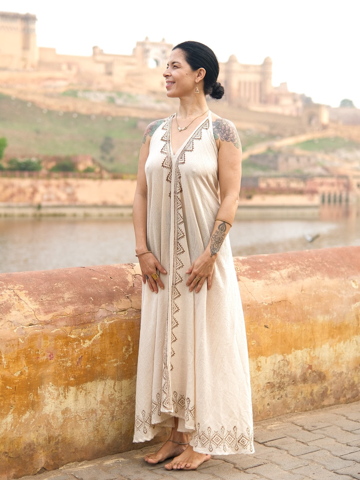 Wearing the Hanvi maxi dress—a sleeveless piece crafted from sustainable cotton and adorned with block print geometric patterns—a woman stands by a river, smiling. Her tattoos are visible on her arms as the dress beautifully complements the old buildings and green hills in the background.