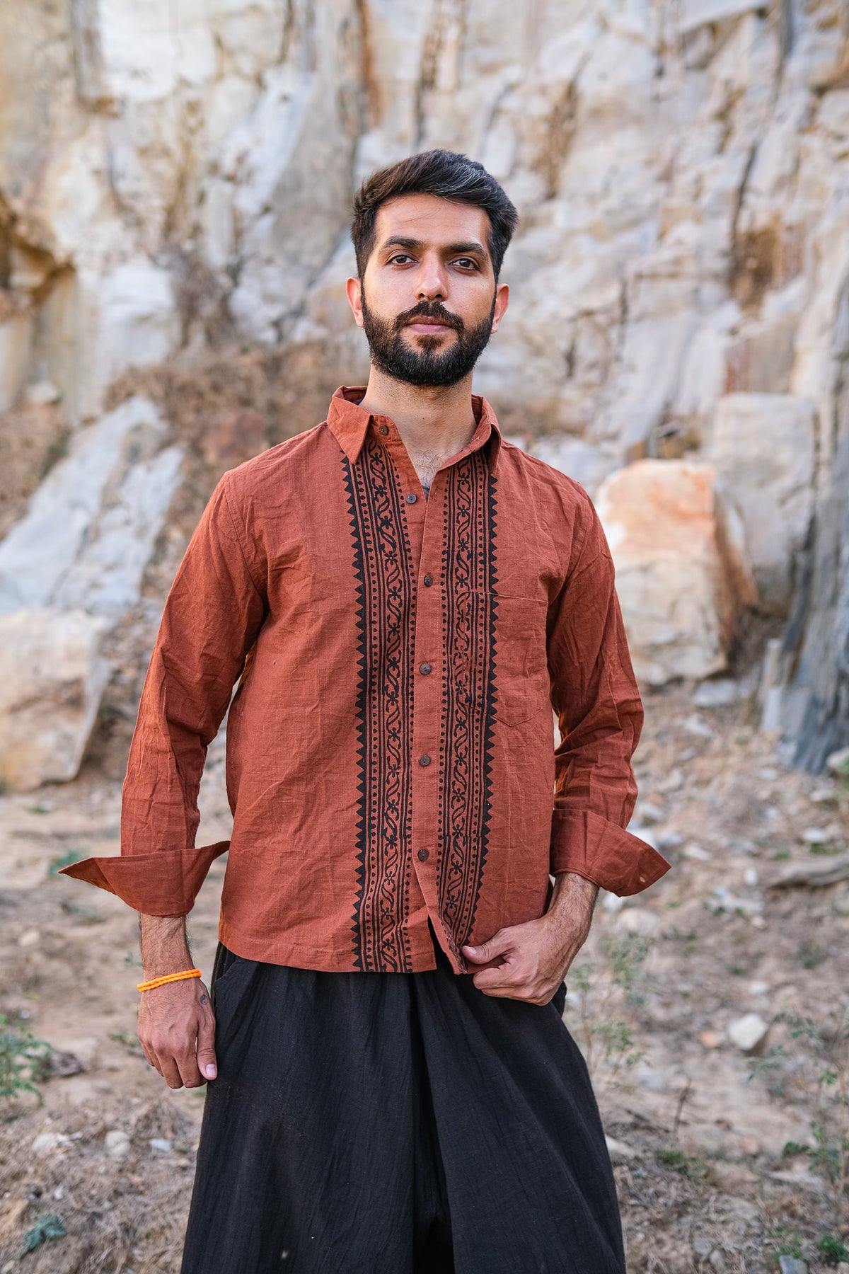 A bearded man confidently stands outdoors wearing the Acharya Shirt Full-sleeve in brown, made from organic cotton with intricate block print designs. His dark pants complement the rugged texture of the rocky background.