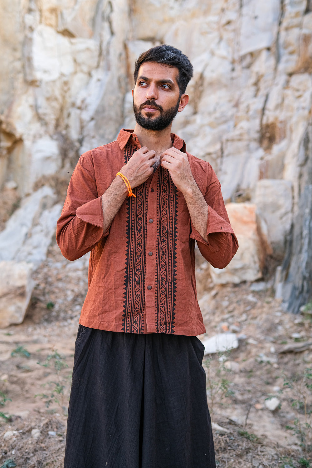 A bearded man adjusts the collar of his brown Acharya Shirt Full-sleeve while standing outdoors in front of rocky terrain. He is also wearing loose black pants.