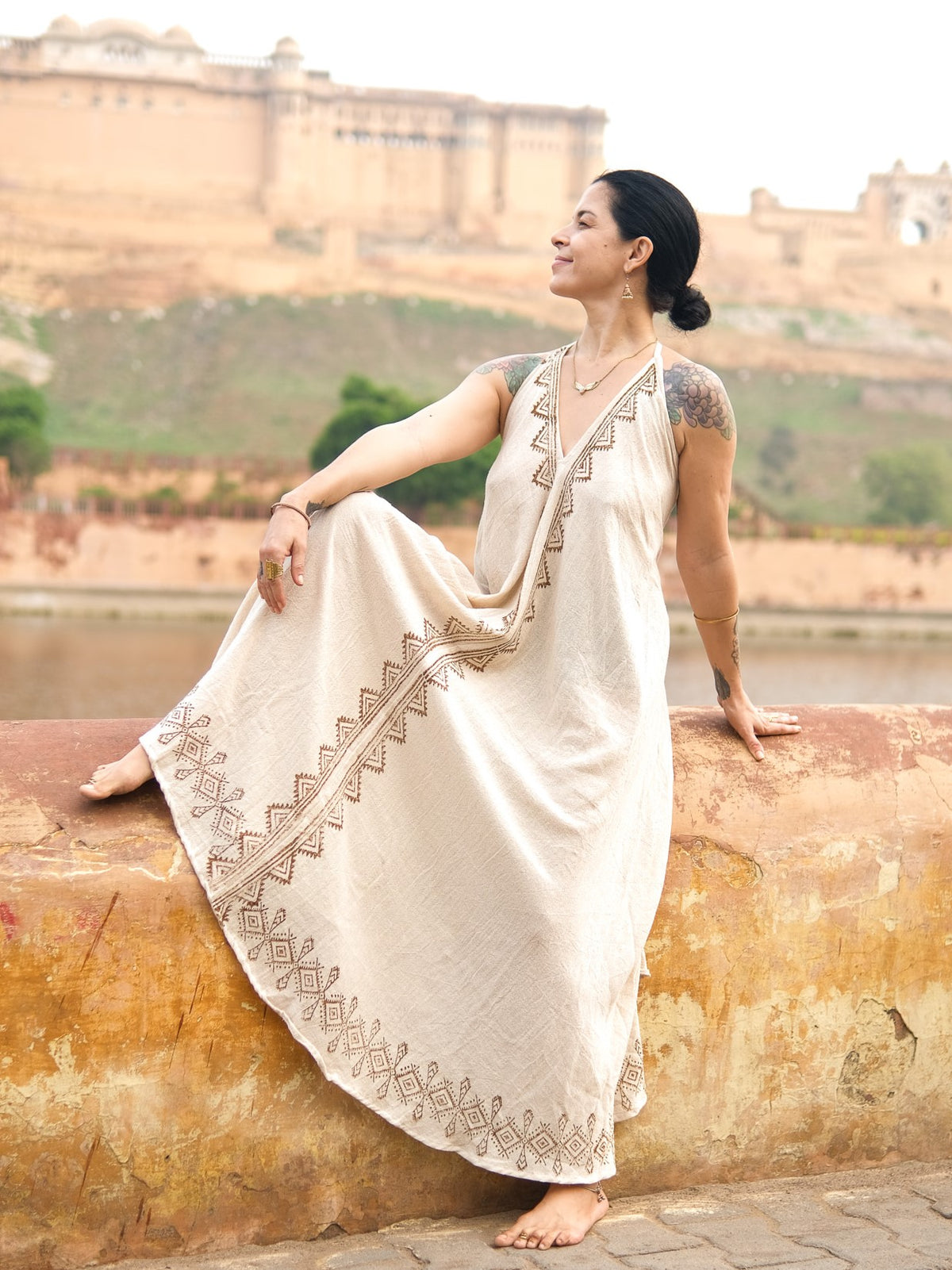 A woman sits barefoot on a ledge, smiling and looking sideways, wearing the Hanvi maxi dress in a cream shade adorned with intricate block print patterns. The sustainable cotton fabric of the dress adds effortless elegance to her relaxed pose, as a large historic fort and lush greenery create a scenic backdrop.