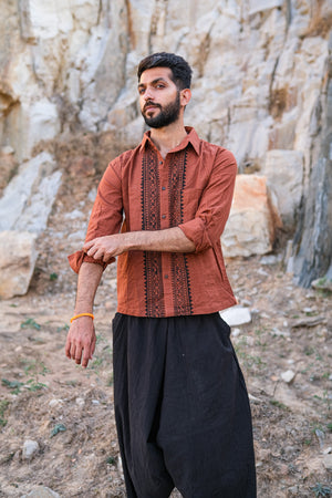 A man wearing the Acharya Shirt Full-sleeve, styled in a brown block print and paired with black pants, rolls up his sleeve while standing outdoors against a rocky background.