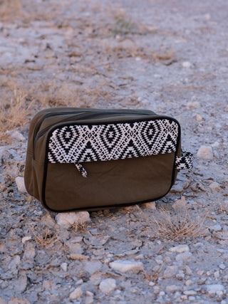 The Vakta Utility Bag boasts ample storage with a green base and chic black and white geometric pattern. It features a zippered compartment, small handle, and sits on a rugged outdoor surface.