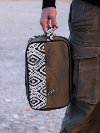 Wearing olive cargo pants, a person holds the Vakta Utility Bag, known for its spacious khaki design and black and white geometric patterned handle. The bags stylish functionality is accentuated against a blurred outdoor backdrop.