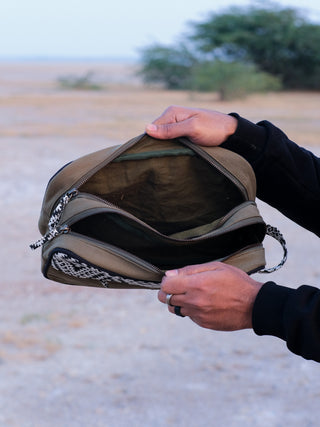 An individual holds an open Vakta Utility Bag, showcasing its spacious two compartments with a cheetah print strap. The outdoor backdrop of dry land and sparse greenery enhances the rugged yet stylish appearance.