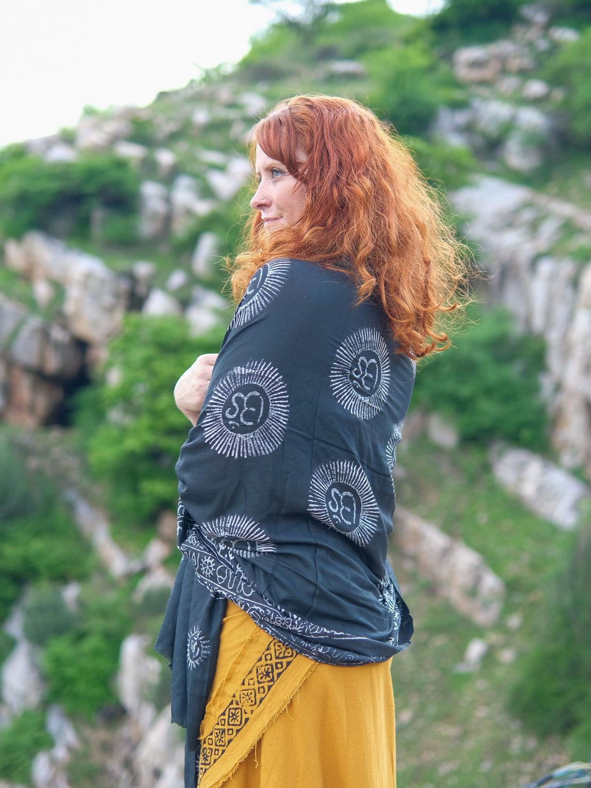 A woman with long red hair stands outdoors, draped in the Mantra Shawl—a luxurious viscose wrap featuring white sunburst patterns—set against a backdrop of green hills and rocky terrain.
