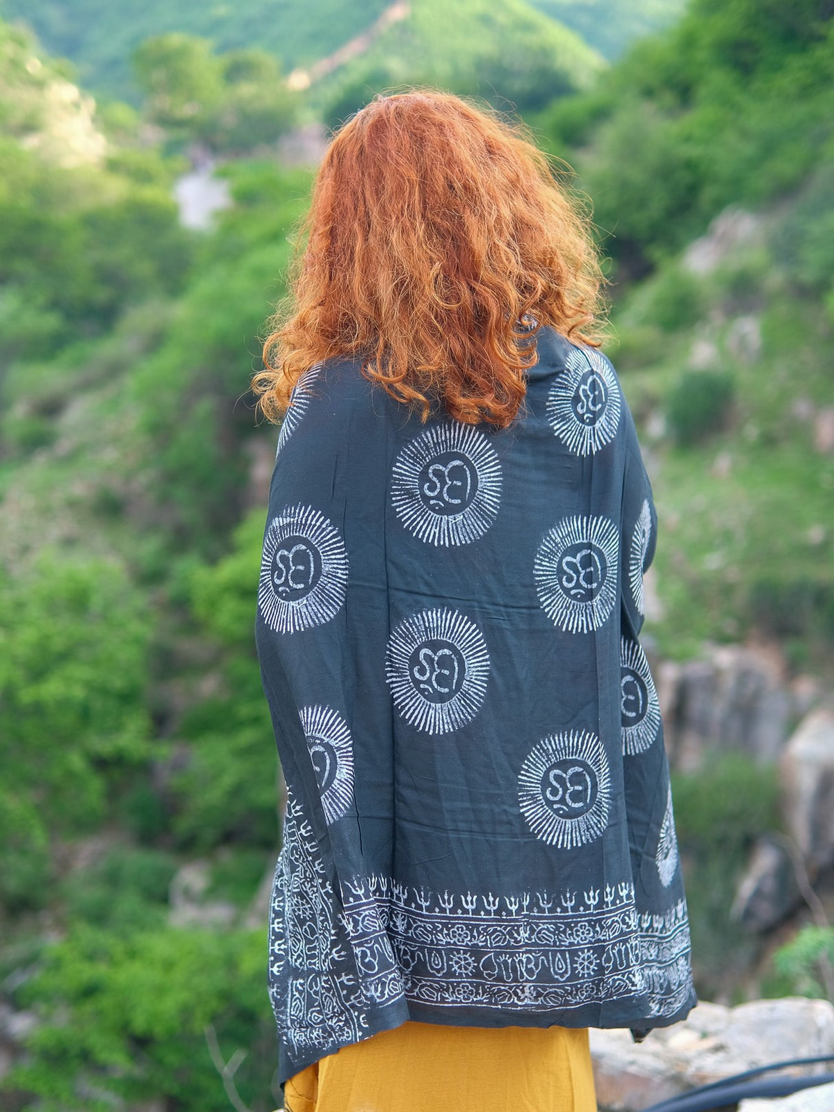 A person with curly red hair, wearing the Mantra Shawl—crafted from luxurious viscose and featuring dark fabric adorned with white patterns—stands outdoors facing lush green hills.