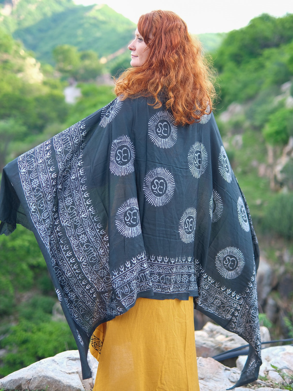 A woman with long red hair, wearing a yellow dress and a luxurious black Mantra Shawl with white patterns, stands outdoors on rocks with a green, mountainous background.
