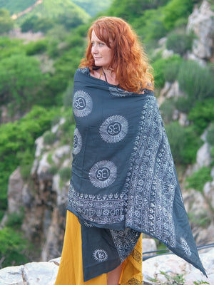 A woman with red hair stands outside, wrapped in the Mantra Shawl decorated with Om symbols, pairing it with a chic bralette and a mustard yellow skirt. The luxurious viscose fabric of the Mantra Shawl glimmers in the sunlight, set against a stunning backdrop of lush green hills and rocky cliffs.