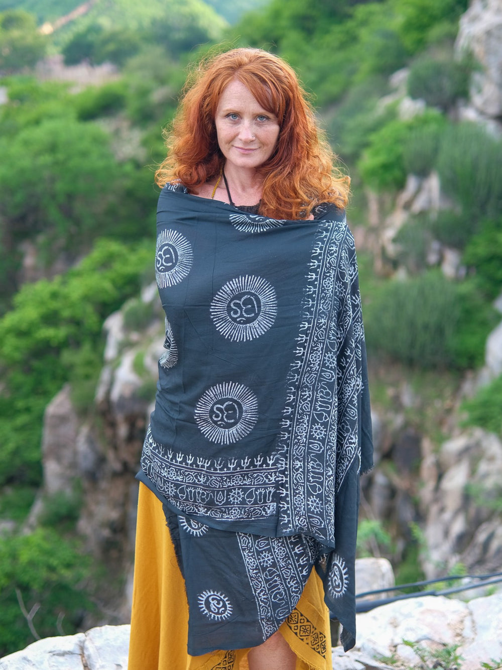A red-haired woman wearing the Mantra Shawl and a yellow skirt stands outdoors before green hills and rocky terrain, gazing at the camera.