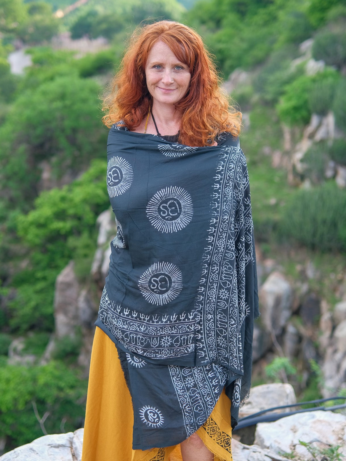A woman with long red hair stands outside, draped in the Mantra Shawl—crafted from luxurious viscose—over a yellow dress, with greenery and rocky cliffs behind her.