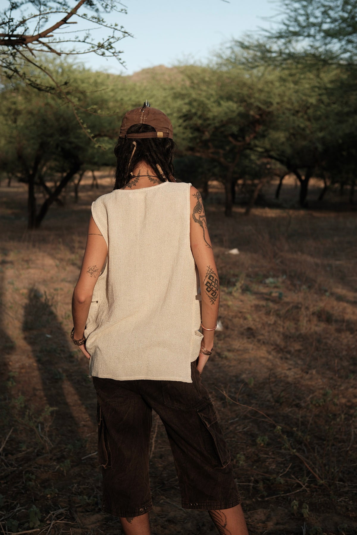 A person with dreadlocks, tattoos, and a brown cap stands outdoors facing away, wearing the Desert Drift Vest in a beige sleeveless style paired with dark shorts—an effortless festival essential—surrounded by trees and dry grass.