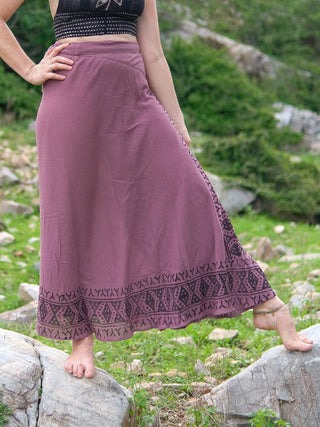 A person stands barefoot on rocks and grass outdoors, wearing the mauve Handloom Wrap Long Skirt with black geometric patterns near the hem and a black crop top, creating a bohemian vibe amid rocky terrain.