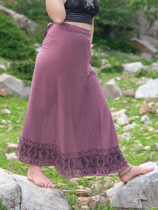A person stands barefoot outdoors on rocks, wearing the Handloom Wrap Long Skirt in mauve with a patterned hem and a black sleeveless top. The grassy background features scattered stones and greenery.