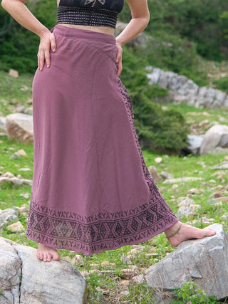A person stands barefoot on rocks outdoors, wearing the Handloom Wrap Long Skirt with patterned borders and a black crop top. One leg is bent and hands are on their waist, surrounded by green grass and rocky terrain.