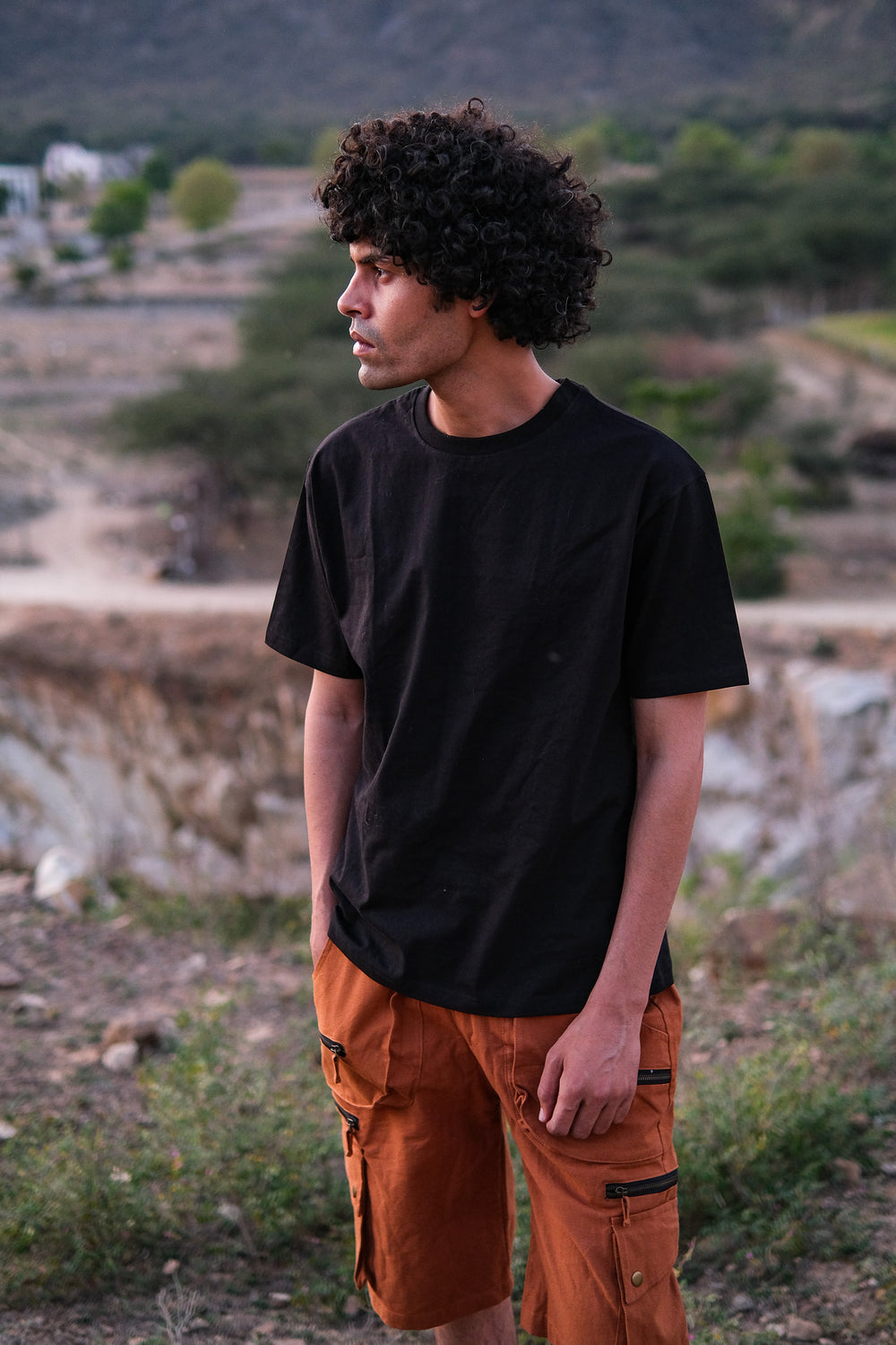 A young man with curly hair, wearing a stylish Alec T-shirt and orange cargo shorts, stands outdoors in a rocky, natural landscape, looking to his left.