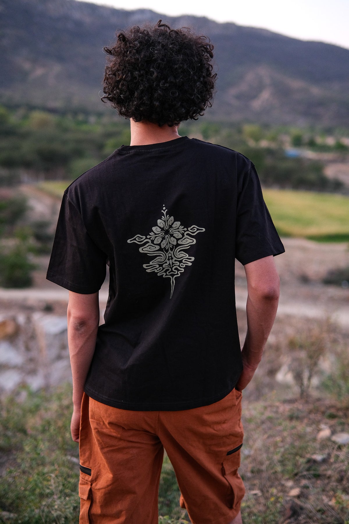 A person with curly hair, wearing a stylish Alec T-shirt crafted from high-quality materials and featuring a floral design on the back, along with orange pants, stands outdoors facing a scenic landscape with mountains and greenery.