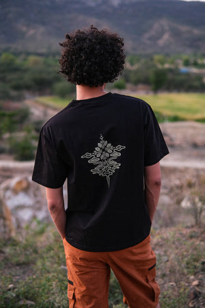 A person with curly hair, seen from behind, stands outdoors wearing the Alec T-shirt—a stylish black tee featuring a floral graphic on the back—paired with orange pants. Nature and mountains are visible in the background.