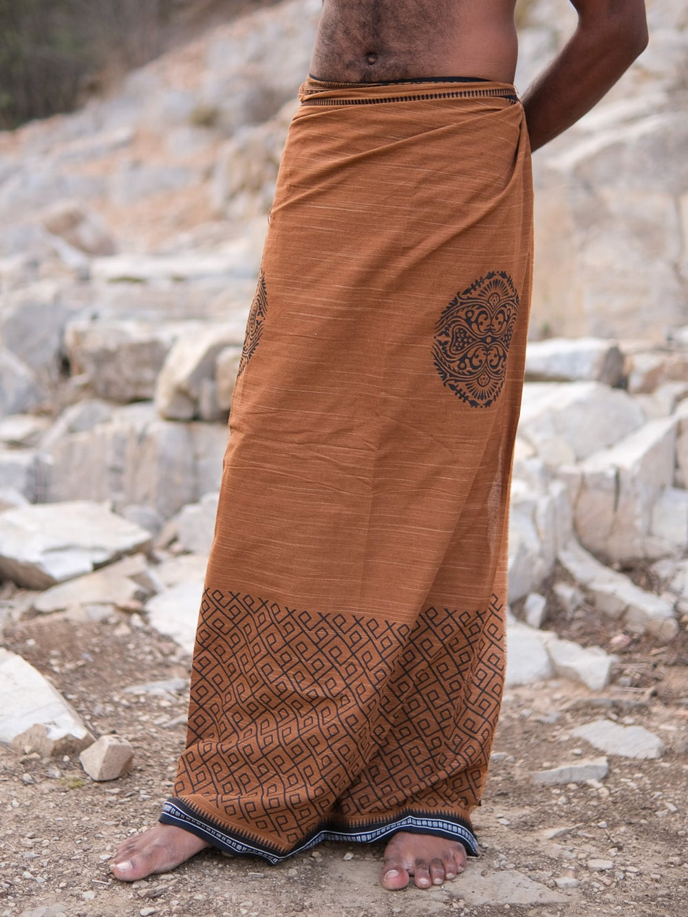 A person standing barefoot on rocky ground, wearing the brown Aastha Dhoti featuring geometric and circular patterns inspired by traditional Rajasthani handblock print. Their upper body and head are out of frame.