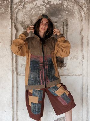 A person stands against a weathered wall, wearing the Achada Utility Jacket and matching shorts featuring patchwork patterns in earthy tones that highlight sustainable fashion, looking at the camera with hands near the hood.