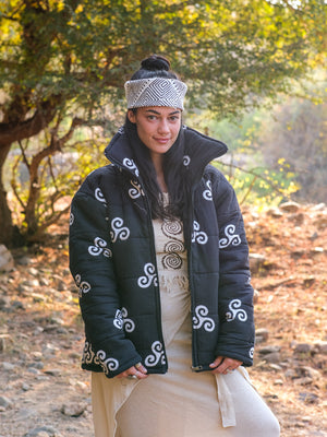 Wearing a patterned headband and the Niruha Jacket—distinguished by its striking black block print with white designs—paired with a beige dress, a person stands outdoors on a sunny day, showcasing Indian fashion amidst trees and sunlight.