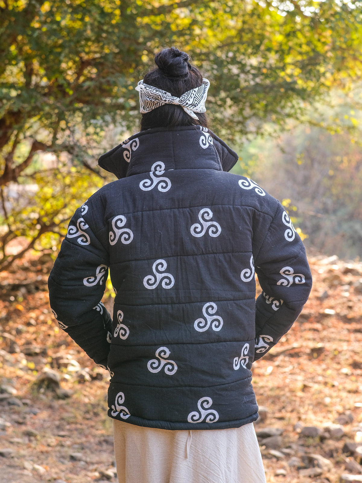 A person stands outdoors with their back to the camera, wearing the Niruha Jacket—a stylish black jacket adorned with white swirl patterns—and a patterned headband, surrounded by trees and sunlight, showcasing a bold blend of Indian fashion inspired by traditional block print jackets.