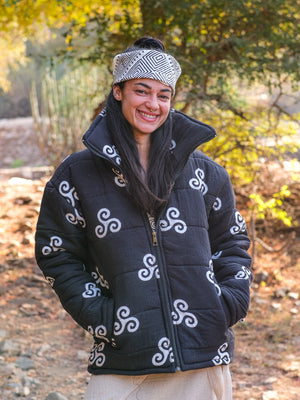 A person wearing a patterned headband and the Niruha Jacket—with black fabric and white spiral designs inspired by Indian fashion—smiles outdoors on a sunlit, wooded path.