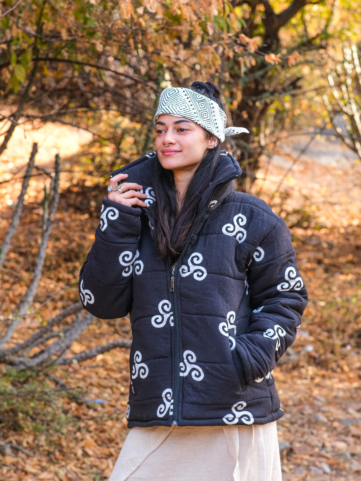 Wearing the Niruha Jacket and a headscarf, a woman stands outdoors in an autumn forest surrounded by yellow and brown leaves and trees, holding something in her hand and smiling slightly.