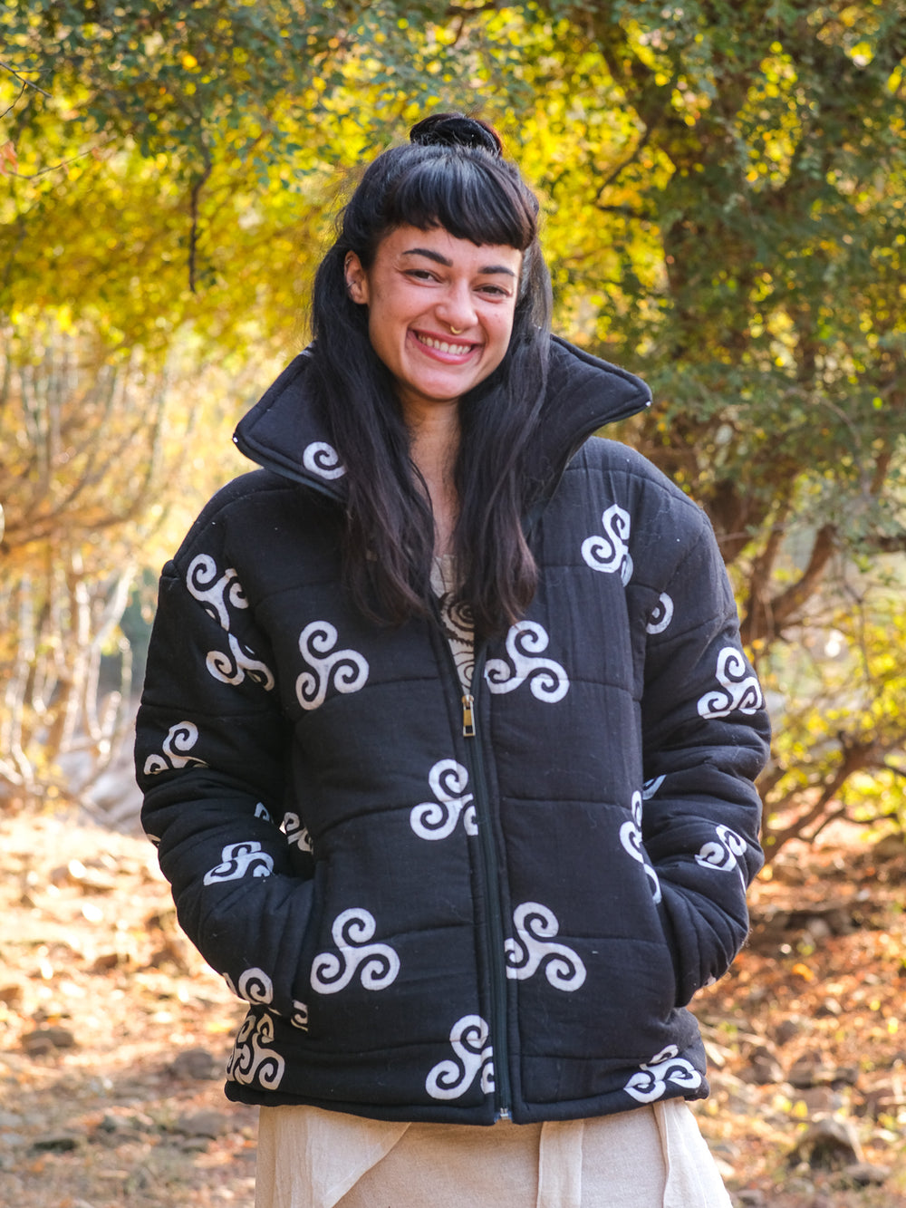 A woman with long dark hair and bangs smiles outdoors, wearing a stylish black Niruha Jacket adorned with white spiral designs inspired by Indian fashion. Trees and sunlight create a vibrant background.