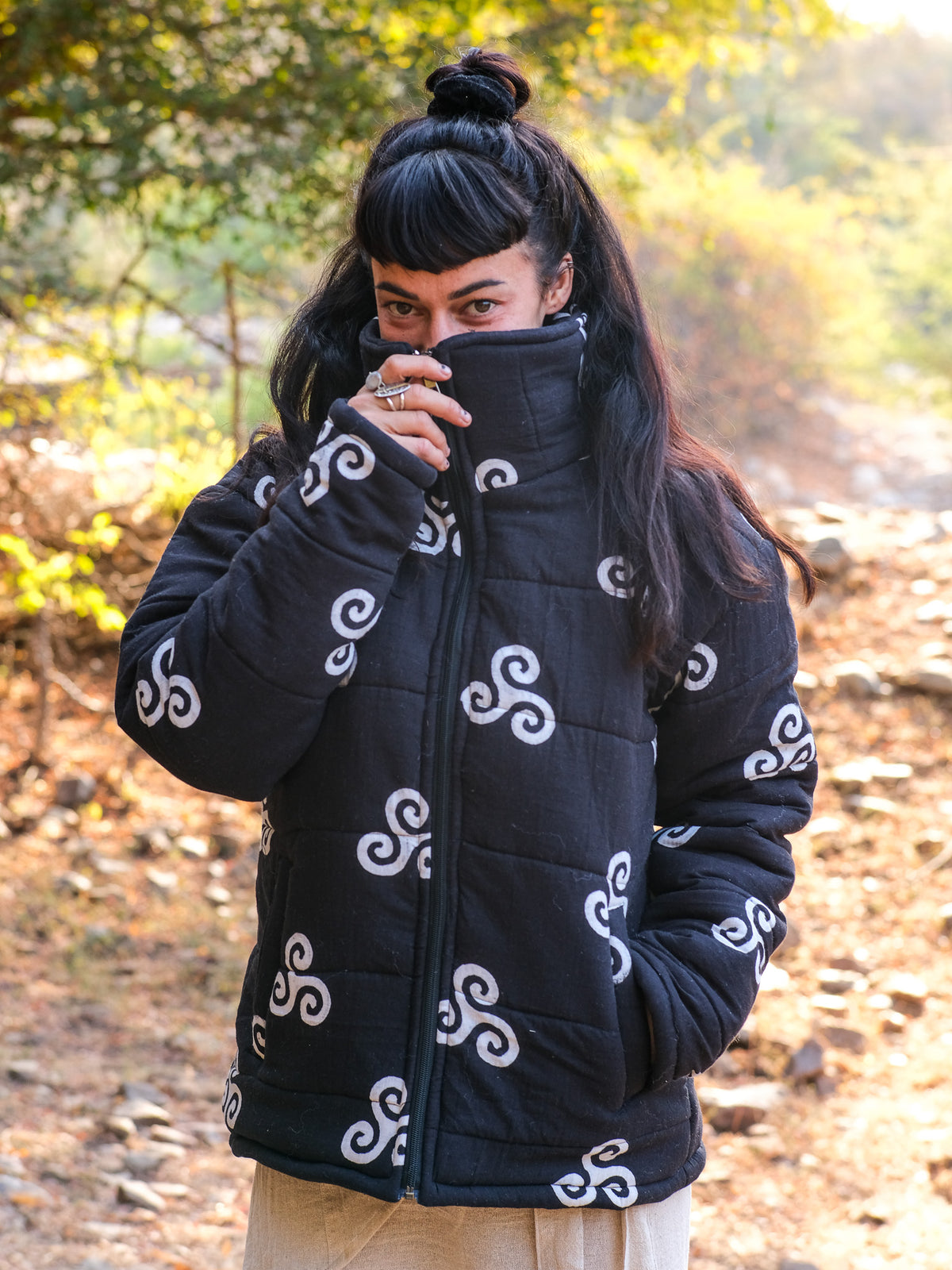 A person with long dark hair styled in a topknot stands outdoors, wearing the Niruha Jacket—a black puffer featuring white swirl designs—and partly covers their face with its high collar. Sunlight filters through the trees behind them, evoking a subtle connection to Indian fashion.