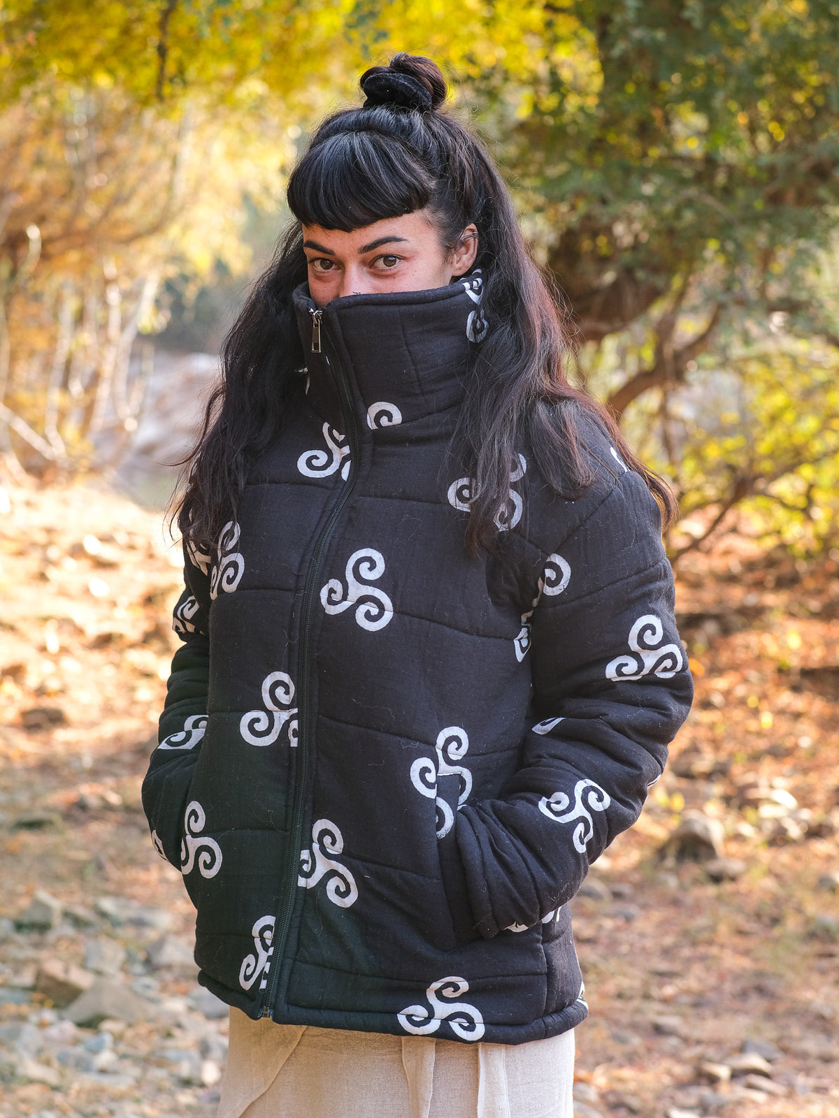 A person with long dark hair and bangs wears the Niruha Jacket in black, which features white swirl patterns, standing outdoors in sunlight with trees and rocks in the background.