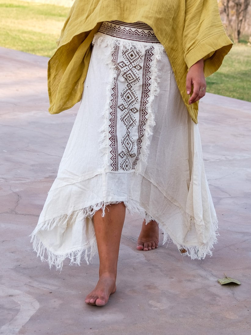 Model walking barefoot on stone paving in the white Nirvighna Skirt with a mustard shawl over her shoulders, showing the brown hand block printed tribal centre panel with fringed edging and the raw-fringed layered asymmetrical cotton hem.