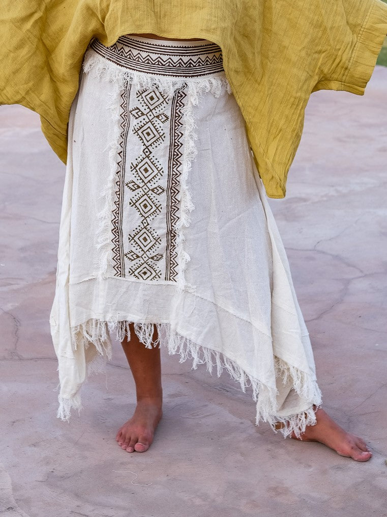 Front view of the white Nirvighna Skirt on stone paving with a mustard shawl draped over the shoulders, showing the full brown hand block printed tribal centre panel with fringed edging and the double raw-fringed asymmetrical cotton hem.