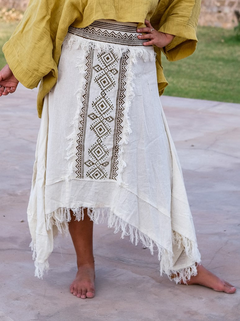 Lower-body view of the white Nirvighna Skirt on stone paving with hands at the hips, showing the brown hand block printed tribal centre panel with fringed edging and the raw-fringed layered asymmetrical cotton hem.