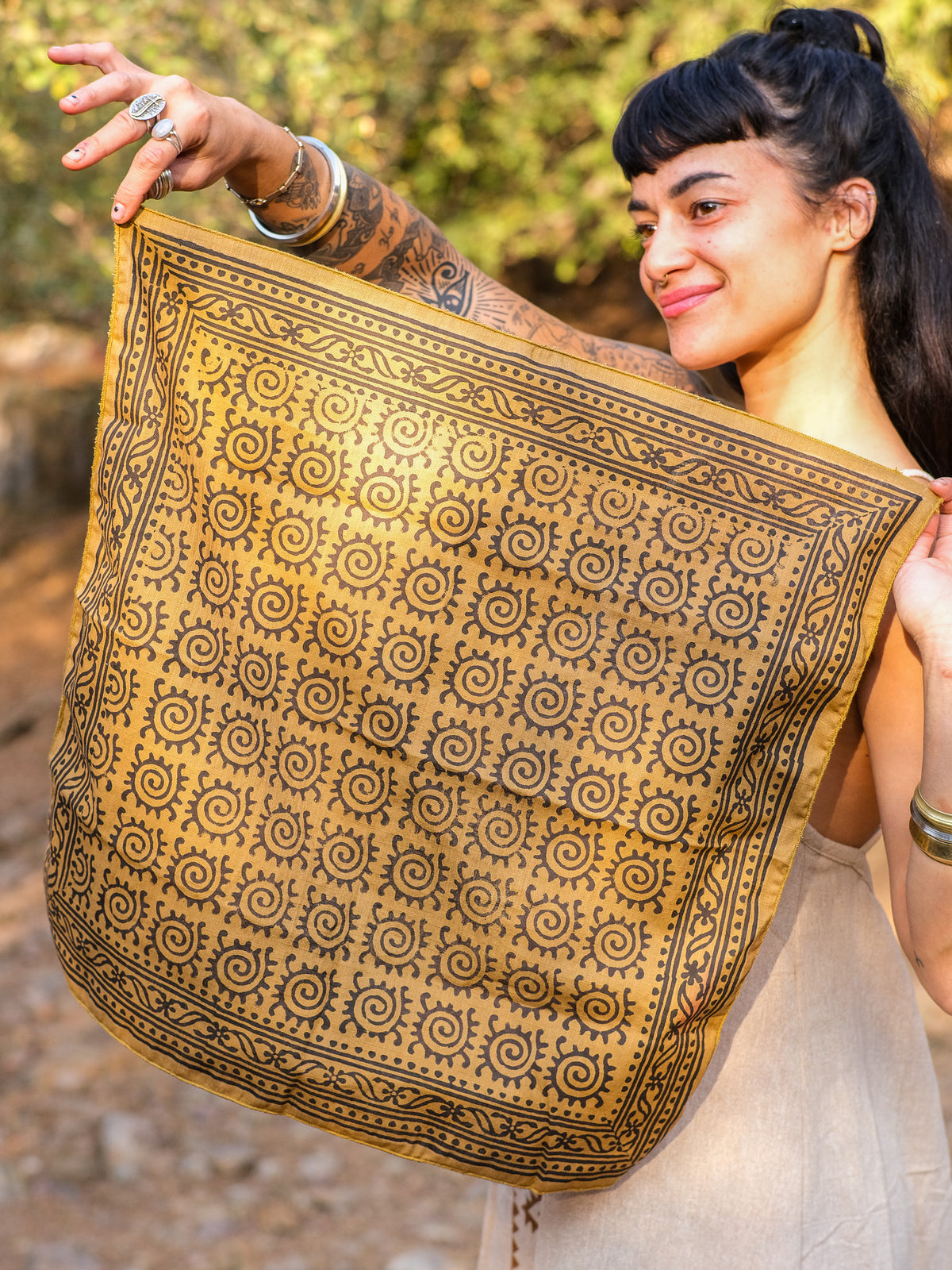 A smiling person with tattoos holds up the green Presh Bandana, a square block-print accessory featuring a repeating black spiral pattern and decorative border, while standing outdoors in natural sunlight.