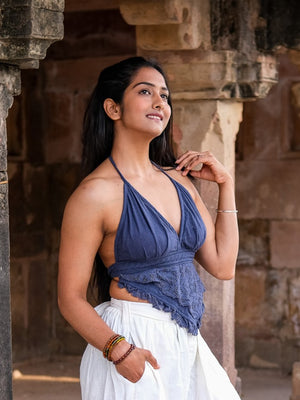 A woman stands outdoors by stone columns, wearing the stylish Titania Crop Top in blue with a white skirt. She smiles gently, looking to the side, with one hand in her hair and the other at her waist.