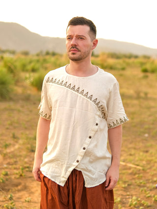 A man wearing the Origin Kurta in natural, featuring tribal embroidery and geometric trim, paired with brown pants, stands outdoors in a grassy sunlit field with hills in the background.