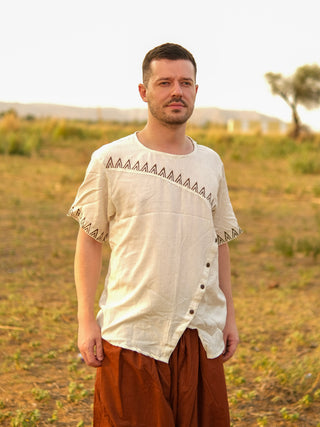 On a sunny day, a man stands outdoors on grassy terrain with a tree and distant hills, wearing the cream-colored Origin Kurta featuring tribal triangle embroidery paired with loose brown pants.