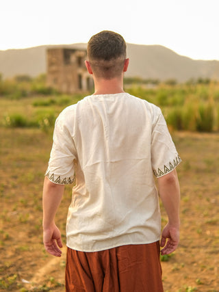 Outdoors against a backdrop of grassy fields, distant hills, and a blurred building, a person stands facing away while wearing the Origin Kurta—a white garment featuring tribal embroidery and patterned sleeves—paired with brown pants.