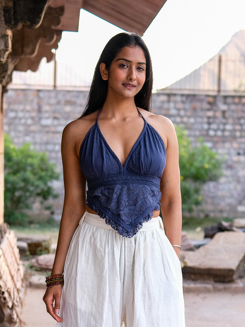 A woman with long dark hair stands outdoors in a blue Titania Crop Top and loose white pants, accessorized with bracelets. She looks relaxed against a stone wall and greenery backdrop.
