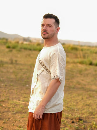 A man with short hair and a beard stands outdoors in a sunlit, grassy field, wearing the Origin Kurta—with geometric patterns in light-colored Rayon Flex fabric—and brown pants. Hills and a distant town are visible in the background.