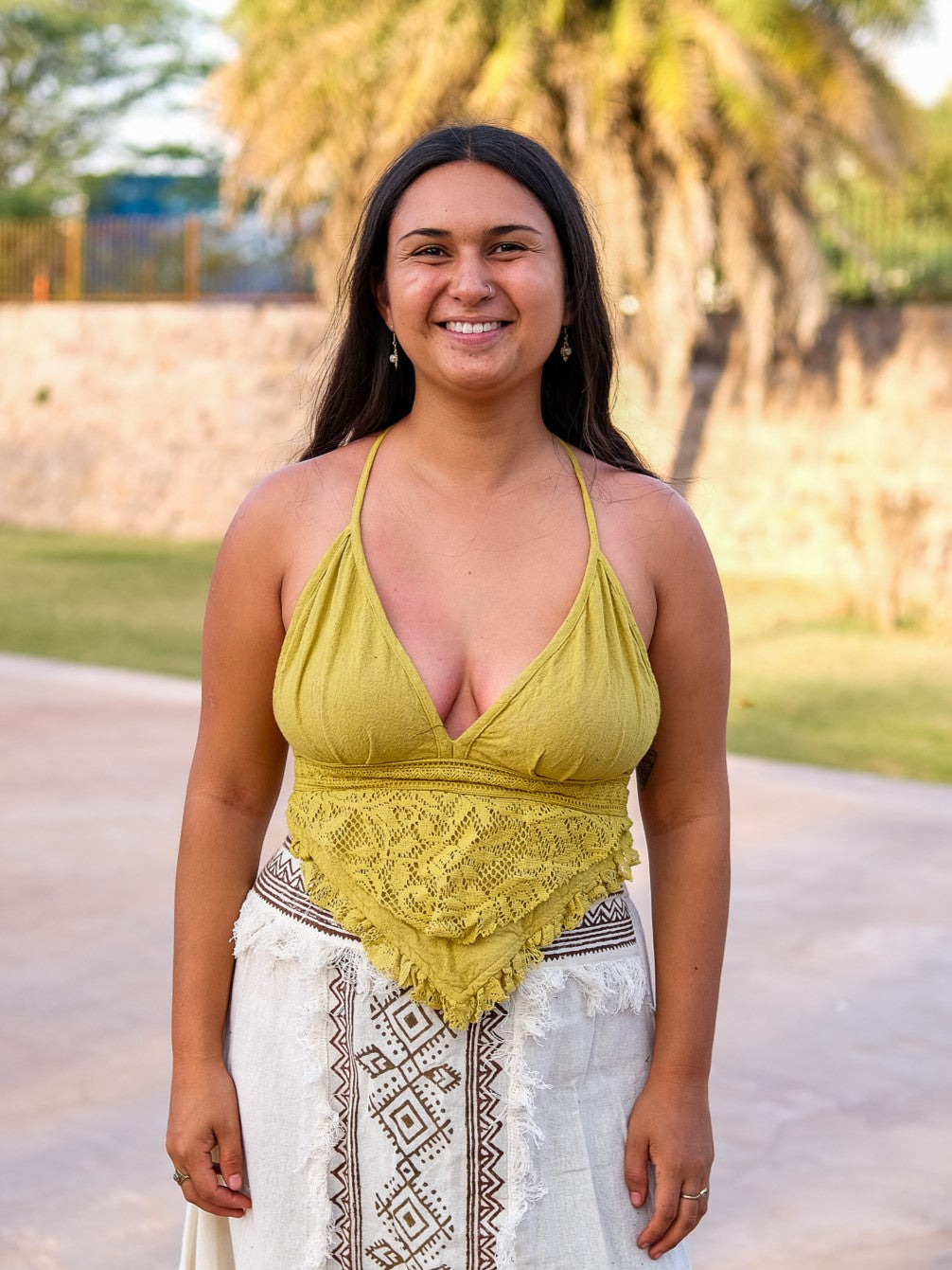 A woman with long dark hair smiles outdoors, wearing a yellow Titania Crop Top and a white geometric-patterned skirt. A palm tree and stone wall create a beautiful backdrop.